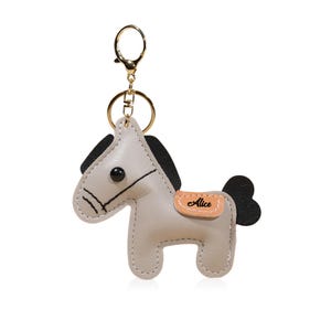 May include: A light gray leather horse keychain with black mane and tail. The keychain has a gold-colored ring and clasp. The horse has a small tan patch with the name "Alice" on it. The horse is a cute, whimsical design.