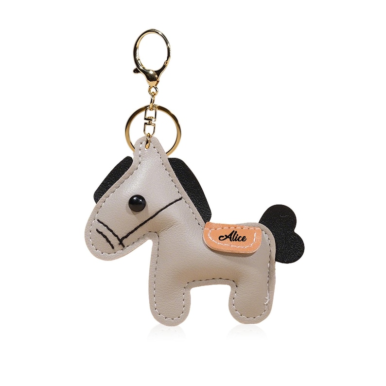 May include: A light gray leather horse keychain with black mane and tail. The keychain has a gold-colored ring and clasp. The horse has a small tan patch with the name "Alice" on it. The horse is a cute, whimsical design.