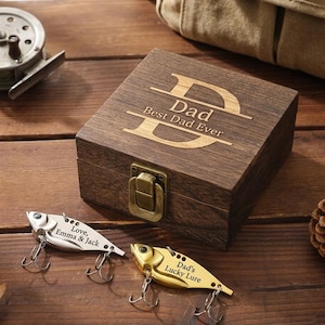 Könnte beinhalten: Eine Holzkiste mit den Worten "Dad Best Dad Ever" auf dem Deckel eingraviert. Zwei Angelköder befinden sich vor der Kiste. Ein Köder ist silberfarben mit den Worten "Love, Emma & Jack" eingraviert. Der andere Köder ist goldfarben mit den Worten "Dad's Lucky Lure" eingraviert.