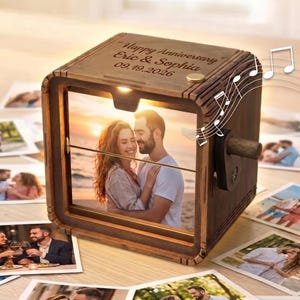 May include: A wooden music box featuring a square frame with a photograph of a couple on a beach. The top is engraved with the text Happy Anniversary Eric & Sophia 09.19.2026. A small wooden crank handle is attached to the side of the dark wood box.