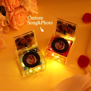 Könnte beinhalten: Zwei transparente Acrylboxen mit Fotos und einem Vinyl-Schallplatten-Design. Die Boxen sind mit Lichterketten beleuchtet und enthalten dekorative Elemente. Der Text "Custom Song & Photo" ist sichtbar. Die Boxen stehen auf einer gelben Oberfläche mit Rosenblättern.