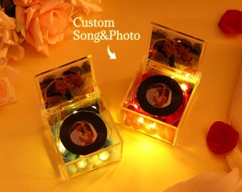 May include: Two clear acrylic boxes with photos and a vinyl record design. The boxes are lit with fairy lights and contain decorative elements. The text "Custom Song & Photo" is visible. The boxes are on a yellow surface with rose petals.
