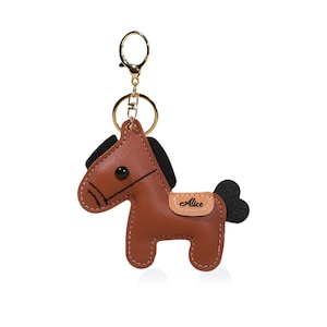 May include: A brown leather horse keychain with black mane and tail. The keychain has a gold-colored clasp and ring. The word "Alice" is printed on a small tan patch on the horse's side. The horse is stitched with white thread.