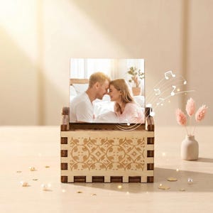 Pode incluir: Uma caixa de música de madeira com uma foto quadrada de um casal no painel frontal. A caixa tem um acabamento em madeira clara com um padrão decorativo cortado a laser na frente. Notas musicais são ilustradas flutuando à direita.