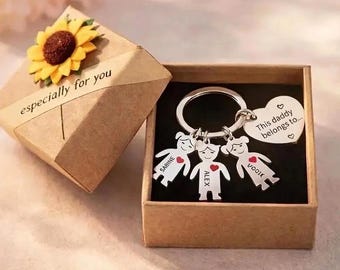 May include: A silver keychain with three figure charms, each with a name and a heart. The keychain is in a brown gift box with the words "This daddy belongs to..." on a heart-shaped charm. The box is next to a gift box with a sunflower.