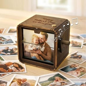 Könnte beinhalten: Eine dunkelbraune Spieluhr aus Holz mit einem quadratischen Rahmen und einem rotierenden Fotodisplay. Auf der Oberseite ist der Text Happy Father's Day, we love you eingraviert. An der Seite befindet sich eine Handkurbel und Musiknoten.