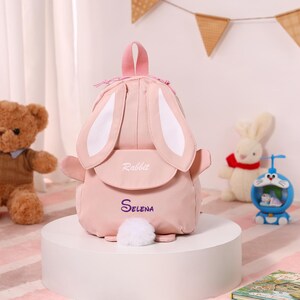 May include: A pink backpack shaped like a rabbit with large ears and a fluffy tail. The backpack has the word "Rabbit" embroidered on the flap and the name "Selena" below. The bag is on a white pedestal.