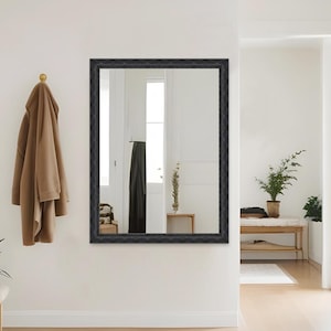 May include: A black framed mirror with an ornate design hangs on a white wall. The mirror reflects a hallway with a coat rack and a plant.
