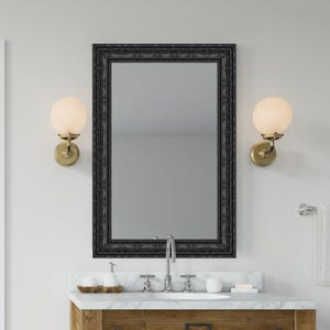 May include: A rectangular black framed mirror hangs above a bathroom vanity. Two sconces with spherical, frosted glass shades flank the mirror. The vanity has a marble countertop and a wooden cabinet.