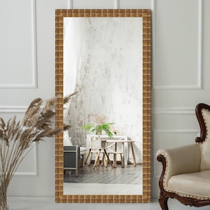 May include: A large, rectangular mirror with a gold-toned frame. The frame has a geometric pattern and is ornate. The mirror is reflecting a room with white walls and wooden furniture.