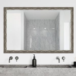 May include: A silver framed bathroom mirror with a beveled edge, hanging above a double sink with two black faucets and a black soap dispenser.