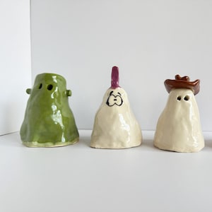May include: Four whimsical ceramic ghost figurines in green, off-white, and red. One ghost has a cowboy hat, another a purple crest, and a third has a cartoon face. The figurines are handmade and suitable for Halloween decor.