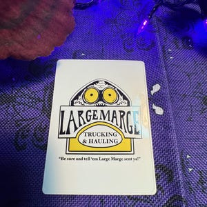 May include: A white rectangular card with the text "LARGEMARGE TRUCKING & HAULING" in black and yellow. The card features a cartoon image of a face with large yellow eyes. The text "Be sure and tell 'em Large Marge sent ya!" is also on the card.