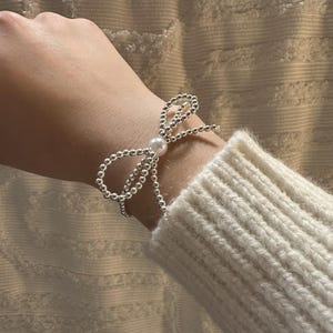 May include: A silver beaded bracelet in a bow design with a single pearl in the center. The bracelet is worn on a wrist, with a white sweater sleeve in the background. The bracelet is made of small, round silver beads.
