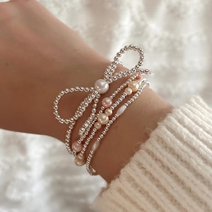 May include: A set of four silver bracelets with pearl accents. The top bracelet features a silver bow with a larger pearl in the center. The other bracelets are layered with smaller pearls in white and pink.