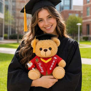May include: A tan plush teddy bear wearing a black graduation cap with a yellow tassel and a red graduation stole. The stole features the white embroidered text Audrey and 2026, along with a small graduation cap icon. The bear holds a white diploma.