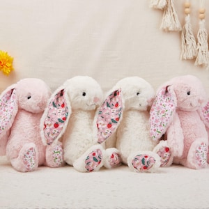 May include: Four plush bunny rabbits in pink and white. The bunnies have long floppy ears and paws with a floral pattern in pink, red, and green. The bunnies are sitting on a white surface.