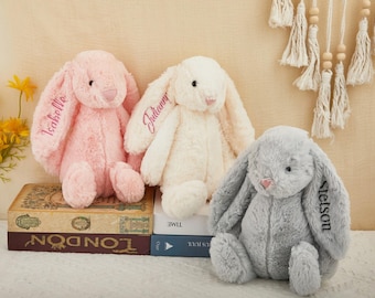 May include: Three plush bunny rabbits in pink, cream, and gray. Each bunny has long ears with embroidered names. The pink bunny has the name "Isabelle", the cream bunny has "Julianny", and the gray bunny has "Stetson".