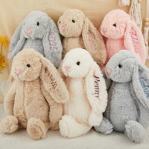 K&ouml;nnte beinhalten: Eine Sammlung von Pl&uuml;schhasen in verschiedenen Pastellfarben, darunter Grau, Rosa, Beige und Wei&szlig;. Jeder Hase hat gestickte Namen auf den Ohren. Die Hasen haben rosa Nasen und sind auf einer cremefarbenen Oberfl&auml;che angeordnet.