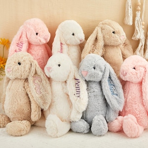 K&ouml;nnte beinhalten: Eine Sammlung von Pl&uuml;schhasen in verschiedenen Pastellfarben, darunter Rosa, Beige und Grau. Jeder Hase hat lange, schlaffe Ohren mit gestickten Namen. Die Hasen sind weich und kuschelig.