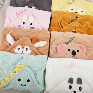 Puede incluir: Una colección de toallas con capucha para bebés en varios colores, cada una con un diseño de cara de animal. Las toallas están bordadas con nombres como "Arlene", "Martin" y "Denise". Los diseños incluyen un conejito, un koala y un dinosaurio.