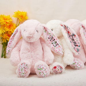 May include: Three plush bunny rabbits in pink and white. The pink bunny has floral patterned ears and feet, with the year "2025" printed on one ear. The white bunny has the name "Emma" printed on one ear. Yellow flowers are in the background.