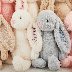 K&ouml;nnte beinhalten: Eine Sammlung von Pl&uuml;schhasen in verschiedenen Pastellfarben, darunter Beige, Rosa und Grau. Jeder Hase hat lange, schlaffe Ohren und gestickte Namen. Die Hasen sind weich und kuschelig, ideal f&uuml;r Kinder.