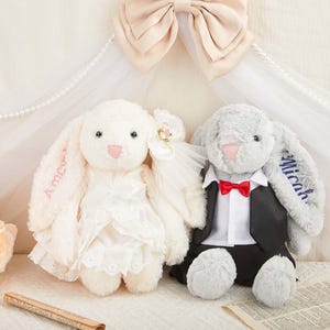 May include: Two plush stuffed rabbits, one cream-colored wearing a white lace dress with a veil and one gray-colored wearing a black tuxedo jacket with a white shirt and a red bow tie. The cream rabbit has the name Amora embroidered on its ear.