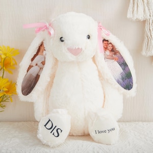 May include: A cream-colored plush rabbit with long ears featuring printed photographs of people. The rabbit has pink ribbons tied at the base of each ear, a pink nose, and black text on its feet that reads DIS and I love you.