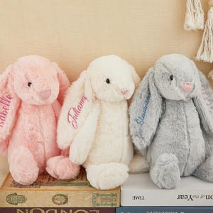 K&ouml;nnte beinhalten: Drei Pl&uuml;schhasen in Rosa, Wei&szlig; und Grau. Jeder Hase hat lange Schlappohren mit gestickten Namen. Der rosa Hase hat den Namen "Isabelle", der wei&szlig;e Hase "Julianny" und der graue Hase "Stevenson".