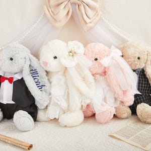 Può includere: Quattro conigli di peluche disposti in fila. Il primo è grigio, indossa uno smoking con papillon rosso e il nome Micah ricamato sull'orecchio. Il secondo è bianco con un velo. Il terzo è rosa con un fiocco bianco. Il quarto è beige con un gilet.