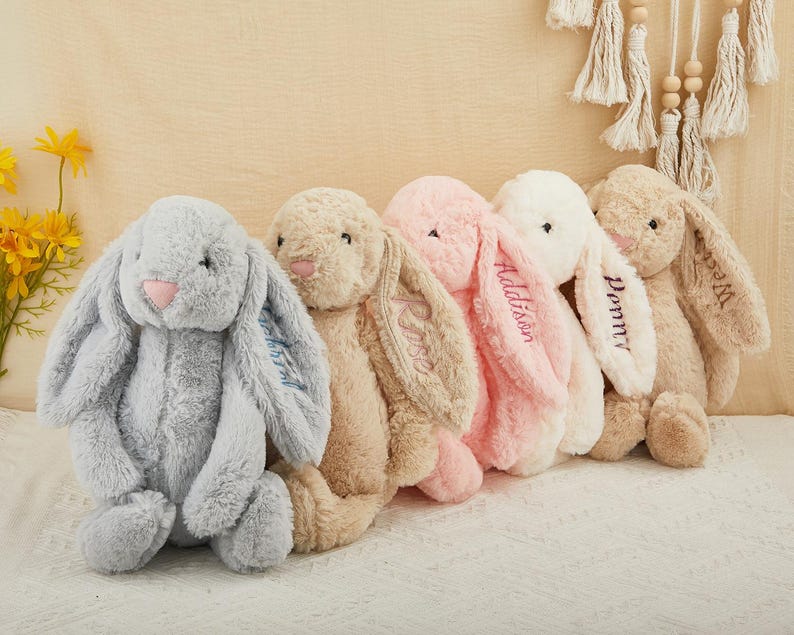 K&ouml;nnte beinhalten: F&uuml;nf Pl&uuml;schhasen in einer Reihe, jeder mit langen, schlaffen Ohren. Die Hasen sind in Grau-, Beige-, Rosa- und Wei&szlig;t&ouml;nen gehalten. Einige Ohren sind mit Namen bestickt. Die Hasen haben rosa Nasen und sitzen aufrecht.