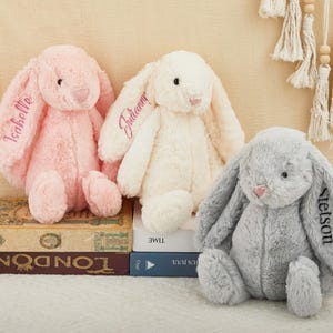 K&ouml;nnte beinhalten: Drei Pl&uuml;schhasen in Rosa, Creme und Grau. Jeder Hase hat lange, schlaffe Ohren und ist mit einem Namen auf einem Ohr bestickt. Die Hasen sitzen auf B&uuml;chern, wobei das Wort "London" sichtbar ist.