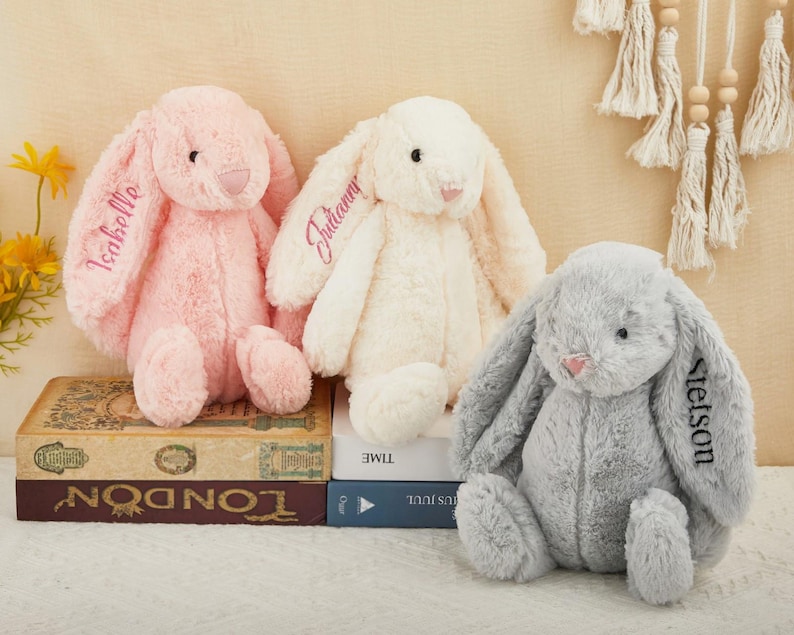 K&ouml;nnte beinhalten: Drei Pl&uuml;schhasen in Rosa, Creme und Grau. Jeder Hase hat lange, schlaffe Ohren und ist mit einem Namen auf einem Ohr bestickt. Die Hasen sitzen auf B&uuml;chern, wobei das Wort "London" sichtbar ist.