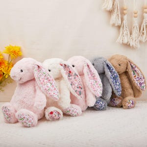 Pode incluir: Cinco coelhos de pelúcia em várias cores: rosa, branco, cinza e castanho. Os coelhos têm orelhas compridas com tecido estampado floral no interior e padrões florais correspondentes nos pés. Estão sobre uma superfície branca.