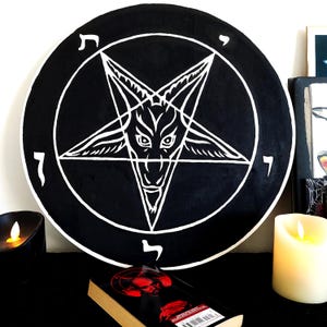 May include: A circular black wall plaque featuring a white hand-painted design of a goat head inside an inverted pentagram. The design is enclosed within a circular border with four Hebrew characters positioned at the top, bottom, left, and right.