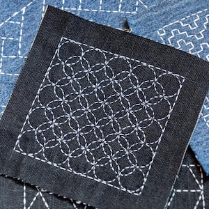 May include: Close-up of several fabric swatches with white stitching. A black square features a repeating circular pattern. Other swatches are denim blue with geometric designs. The stitching creates a textured, handcrafted appearance.