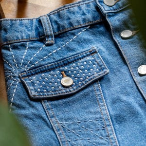 May include: Close-up of a light blue denim jacket with white stitching details. The jacket features a pocket with a button closure and decorative stitching. Silver buttons secure the front.