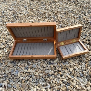 May include: Two open wooden fly fishing boxes with gray foam inserts and small hooks. The boxes are rectangular with a hinged lid and a brass clasp. The boxes are different sizes and are sitting on a bed of small rocks.