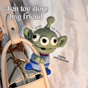 Peut inclure: Un ami de sac Alien Toy Story crochet&eacute; en vert, blanc, bleu et violet. L'alien serre une poign&eacute;e de sac &agrave; main beige. Le texte sur l'image se lit "alien toy story bag friend" et "hugging your bag handle".