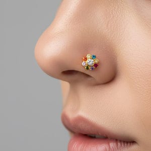Peut inclure: Un bijou de nez en or composé d'une pierre centrale ronde et transparente entourée d'un groupe de petites pierres précieuses multicolores, notamment rouges, bleues, jaunes, orange, violettes et roses, formant un motif floral circulaire.