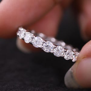 May include: An eternity ring with a continuous line of round diamonds. The ring is held between fingers against a dark backdrop, highlighting the sparkle of the gemstones. The diamonds are set closely together, forming a seamless band.