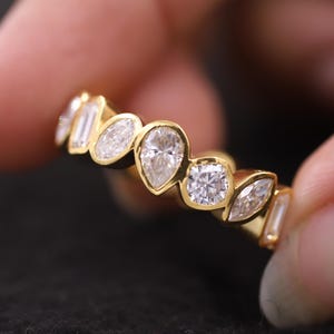 May include: A gold ring with a series of clear gemstones in various shapes. The gemstones include pear, oval, and square cuts. The ring is held between two fingers against a dark background.