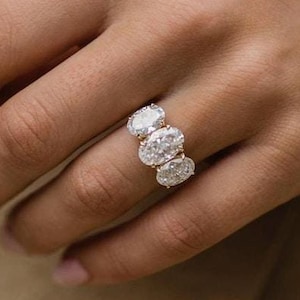May include: A gold ring with three oval-cut diamonds. The ring is set on a woman's hand with a neutral background. The diamonds are clear and sparkling, and the gold band is polished.