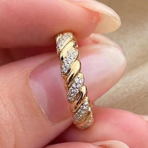 May include: A gold ring featuring a twisted rope design with alternating sections of polished gold and rows of small, clear, sparkling stones embedded along the spiral pattern, creating a textured and reflective appearance on the band.