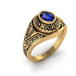 Peut inclure: Une bague dorée ornée d'une pierre précieuse bleue. La bague porte l'inscription "MICHIGAN" et l'année "2021". La monture de la bague présente un motif détaillé.