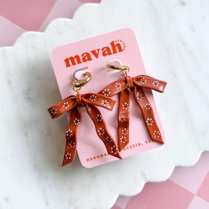 May include: A pair of bow-shaped earrings with a terracotta color and white floral pattern. The earrings are attached to gold hoops and displayed on a pink card with the text "mavah design" and "HANDMADE AUSTIN, TEX".