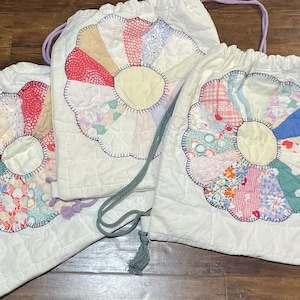 Può includere: Tre sacchetti di stoffa bianca con coulisse e design trapuntato. Ogni sacchetto presenta un diverso design floreale in patchwork colorato al centro.