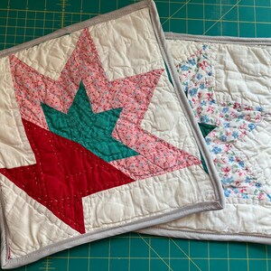May include: Two square quilted potholders with a star pattern. The top potholder features a large star with red, teal, and pink floral fabric on a white background. The second potholder has a similar star design with blue and pink floral fabric.