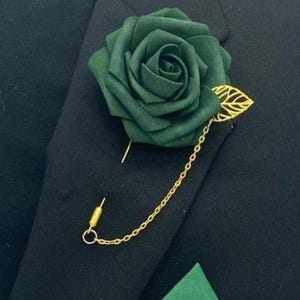 May include: A dark green rose lapel pin with a gold-colored leaf and chain detail. The fabric rose is attached to a gold pin, and the chain connects to a small gold loop. The pin is displayed on a black blazer.